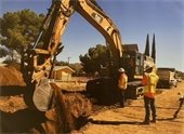 Photo of yellow backhoe and men digging a trench in the street to install new sewer pipeline.